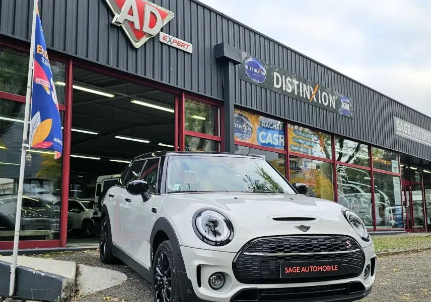 Vue 3/4 avant d'une MINI Cooper S Clubman blanche avec toit noir, garée devant un garage Sage Automobile.