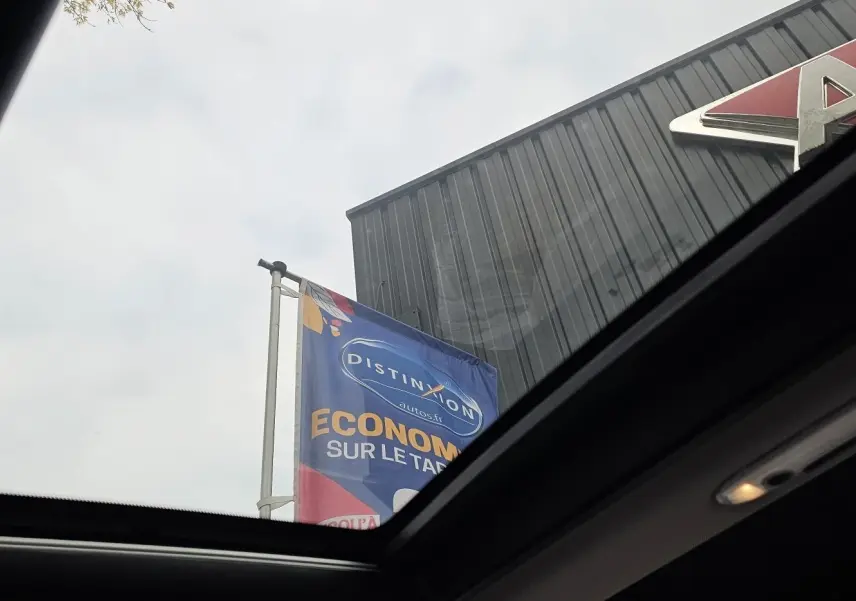 Vue partielle par le toit ouvrant d'un MINI Cooper S Clubman grise 2022, avec ciel nuageux et enseigne de concession visible.