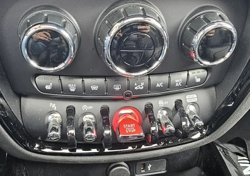 Gros plan sur la console centrale du MINI Clubman grise, mettant en valeur les commandes chromées et le bouton rouge Start/Stop.