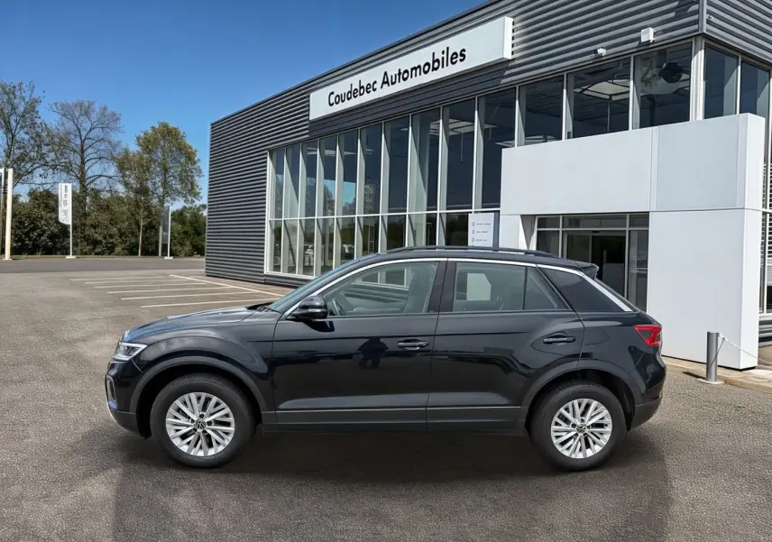 Vue de profil côté gauche d’un Volkswagen T-Roc noir 2025 garé devant un bâtiment de concession automobile.
