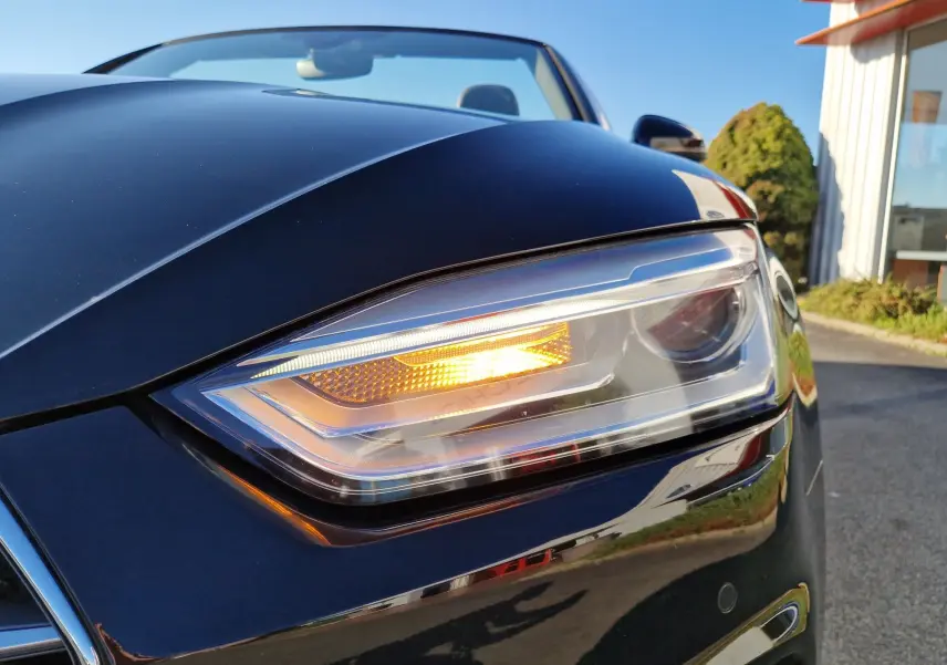 Gros plan sur le phare avant droit allumé d'une Audi A5 Cabriolet noire, soulignant ses lignes élégantes et modernes.