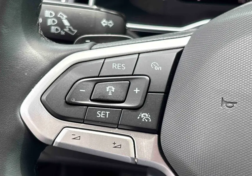 Gros plan sur les commandes multifonctions à gauche du volant en cuir du Volkswagen Taigo blanc, avec boutons de régulateur de vitesse.