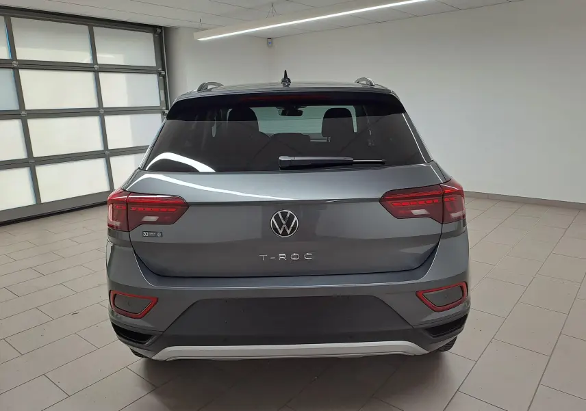 Vue arrière d'un Volkswagen T-Roc gris Indium 2024 avec feux LED et caméra de recul dans un garage lumineux.