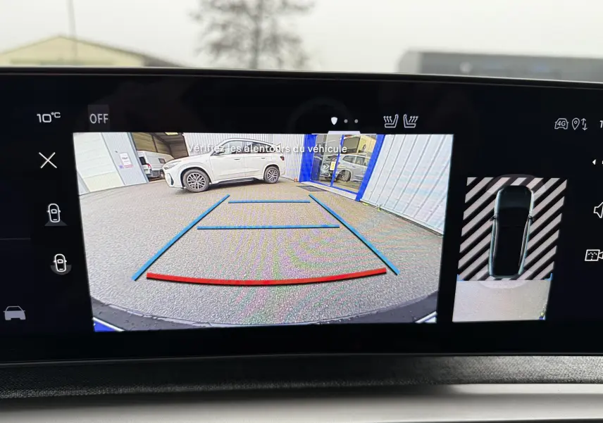 Vue caméra de recul montrant un SUV blanc Okenite Peugeot 3008 hybride garé près d'un bâtiment avec toit noir visible.