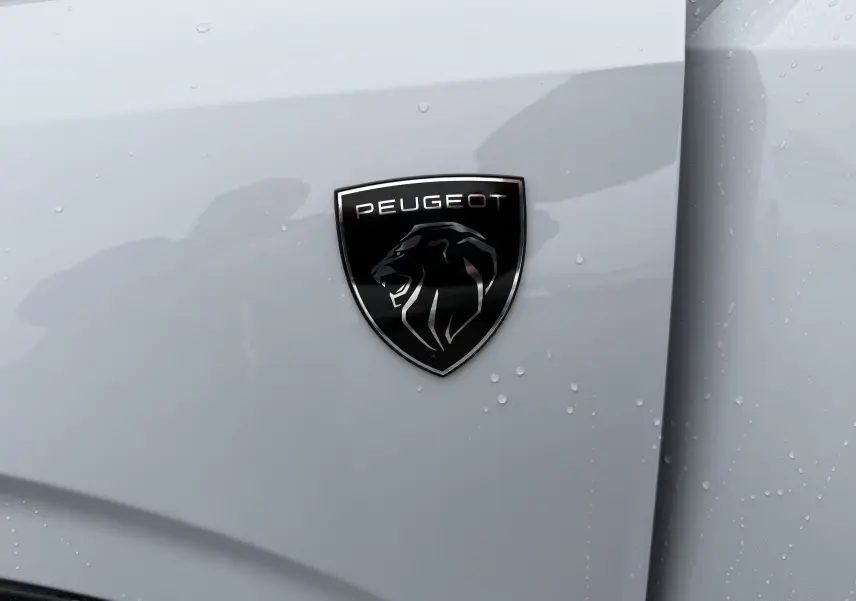 Gros plan sur l'écusson noir Peugeot sur la carrosserie blanche Okenite du SUV 3008 Hybrid 2025.
