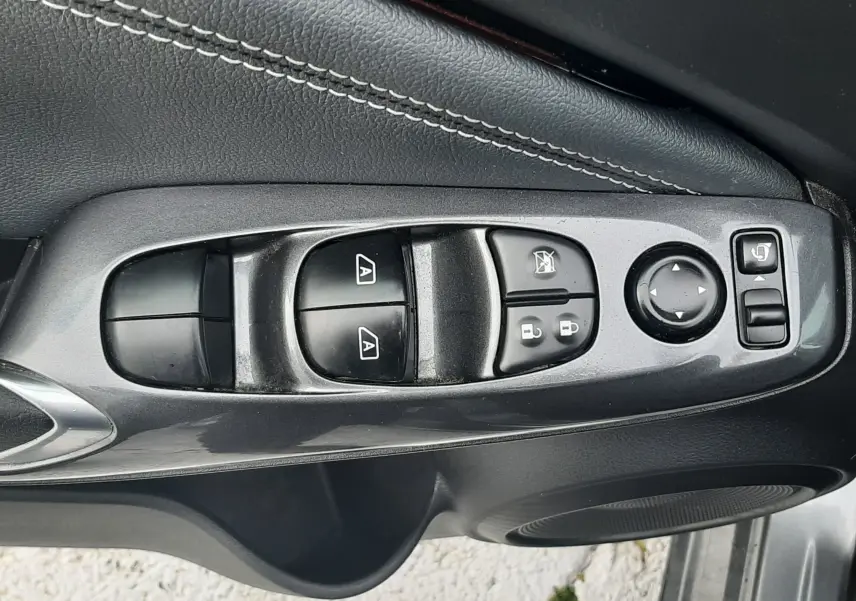 Gros plan sur la commande des vitres et rétroviseurs côté conducteur du Nissan Juke gris, avec surpiqûres blanches visibles.