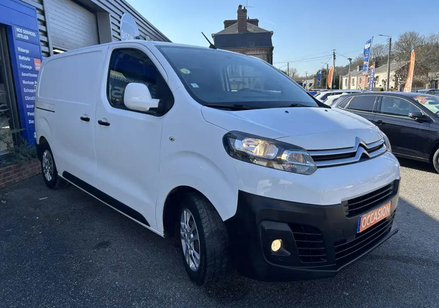 Vue 3/4 avant droite d'un Citroën Jumpy blanc avec attelage, stationné devant un garage en extérieur.
