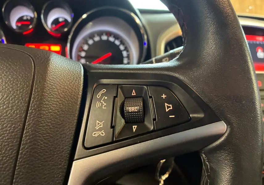 Gros plan sur les commandes multifonctions au volant d'une Opel Astra gris, avec tableau de bord et compteurs en arrière-plan flous.