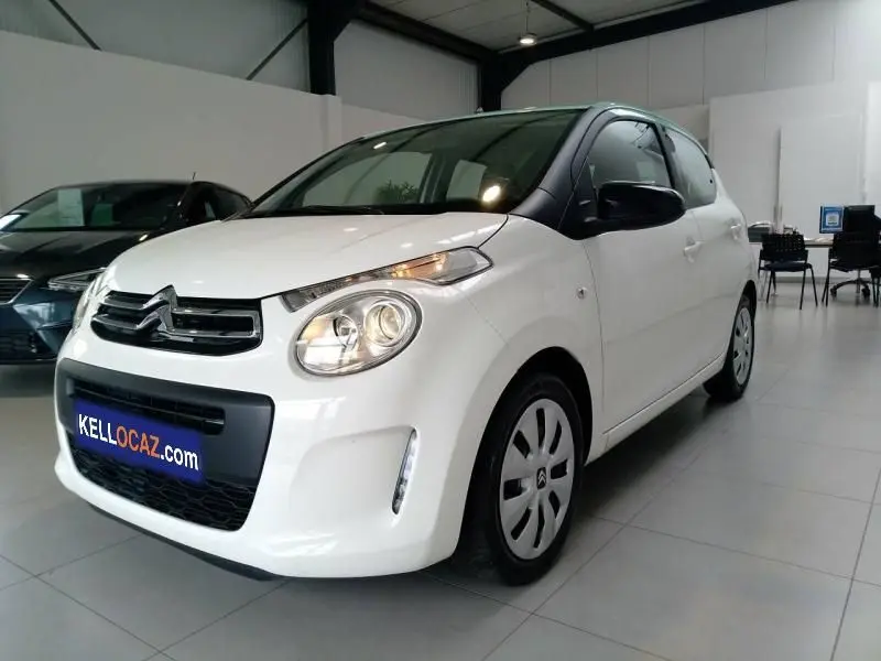 Citroën C1 blanche vue 3/4 avant droit en intérieur avec phares allumés et enjoliveurs noirs sur jantes.