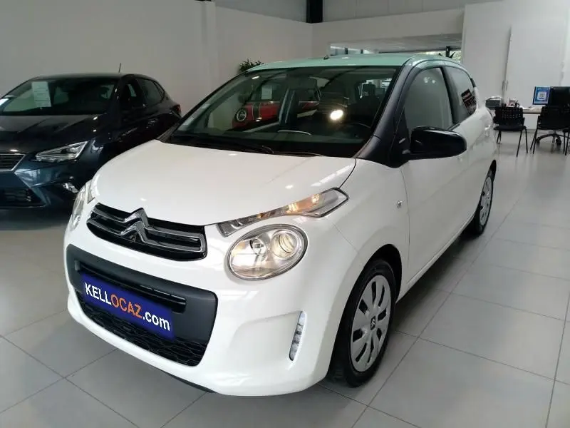 Citroën C1 blanc vue 3/4 avant droit en intérieur, avec phares allumés et enjoliveurs sur jantes acier.