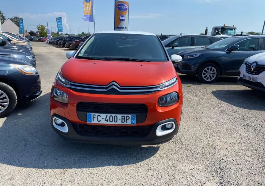 Vue de face d'une Citroën C3 orange avec toit blanc et feux avant distinctifs sur un parking extérieur.