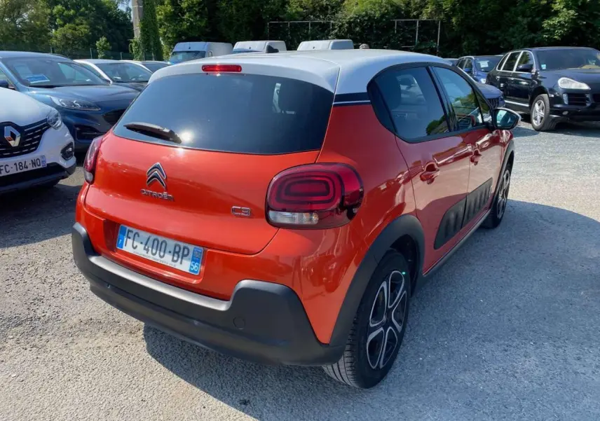 Vue 3/4 arrière droite d'une Citroën C3 orange avec toit blanc et protections latérales noires sur parking.