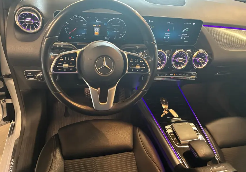 Vue intérieure centrée sur le volant et la console d'une Mercedes Classe B 200D gris, avec éclairage d'ambiance violet.