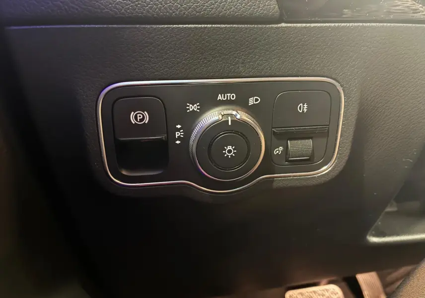 Gros plan sur le panneau de commande des phares et du frein à main électrique dans l’habitacle gris-noir d’une Mercedes Classe B 200D Progressive Line.