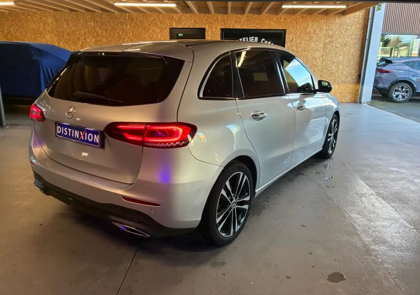 Mercedes Classe B 200D Progressive Line gris vue 3/4 arrière droit, feux arrière allumés en intérieur d'atelier.