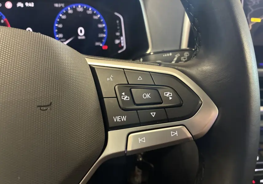 Gros plan sur les commandes au volant noir du Volkswagen T-Roc 2.0 TDI 150 DSG7, avec tableau de bord numérique en arrière-plan.