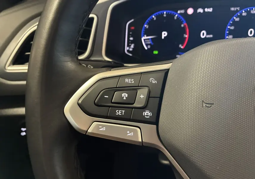 Gros plan sur le côté gauche du volant noir du Volkswagen T-Roc 2025, avec commandes de régulateur et tableau de bord numérique.