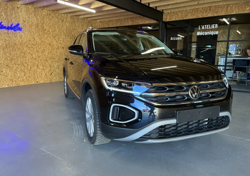 Volkswagen T-Roc noir 2025 en 3/4 avant droit, avec calandre chromée et phares LED, stationné en intérieur.