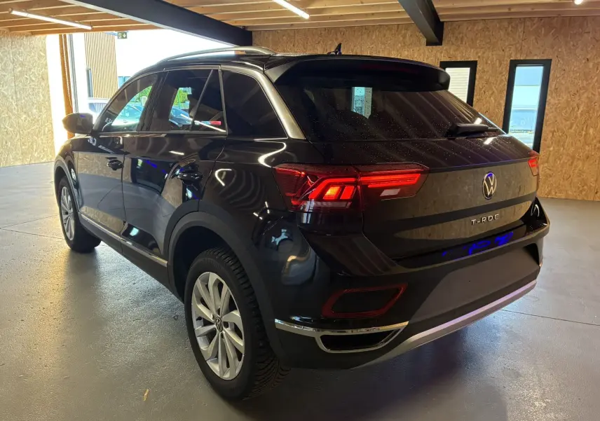 Vue 3/4 arrière droite du Volkswagen T-Roc noir 2025 avec feux arrière allumés dans un garage.