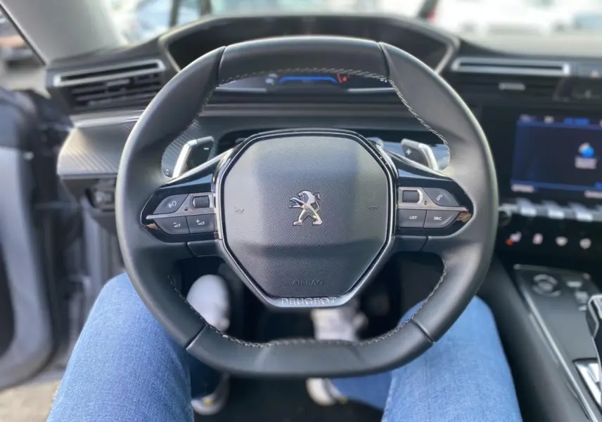 Vue rapprochée du volant compact noir du Peugeot 508 SW 2021, avec tableau de bord numérique en arrière-plan flou.