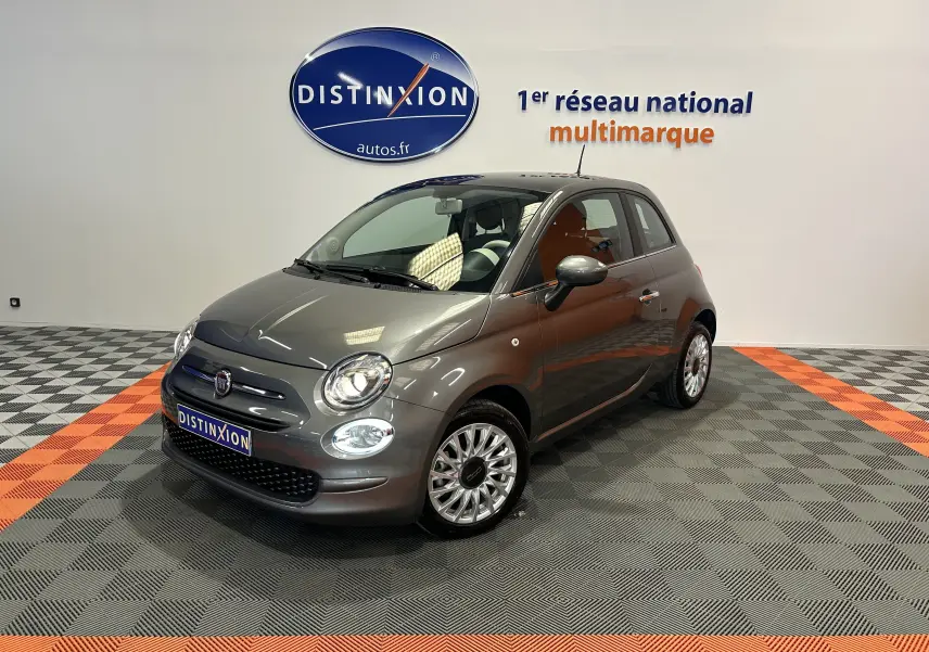 Vue 3/4 avant d'une Fiat 500 grise avec toit panoramique, dans un showroom au sol gris et orange.
