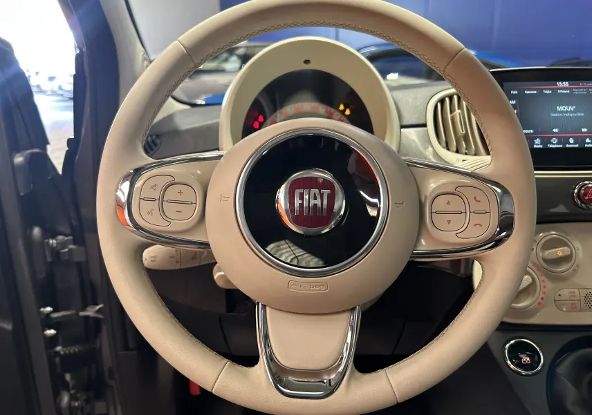 Vue rapprochée du volant beige de la FIAT 500 70 Hybride BSG DolceVita avec tableau de bord et écran tactile visibles.