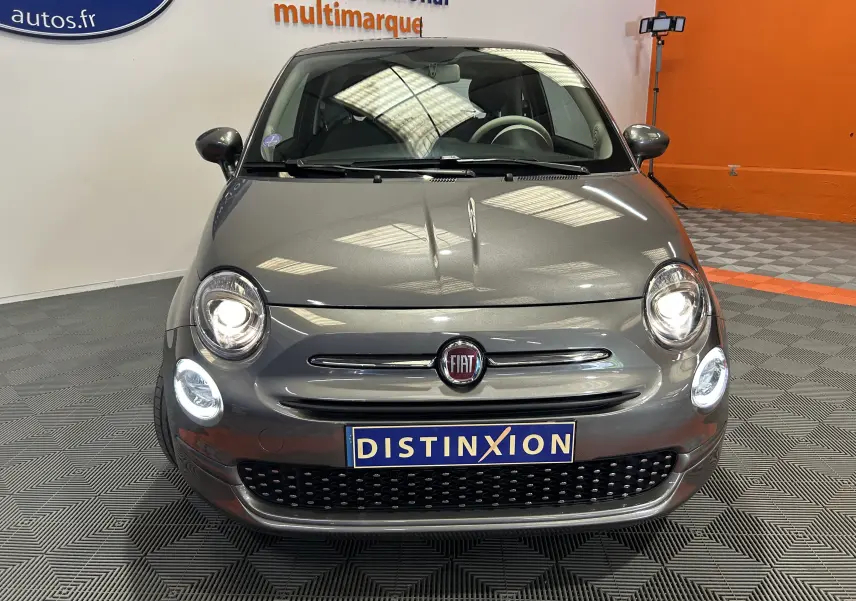 Vue avant d'une FIAT 500 grise 2022 avec phares allumés et toit panoramique visible en intérieur showroom.