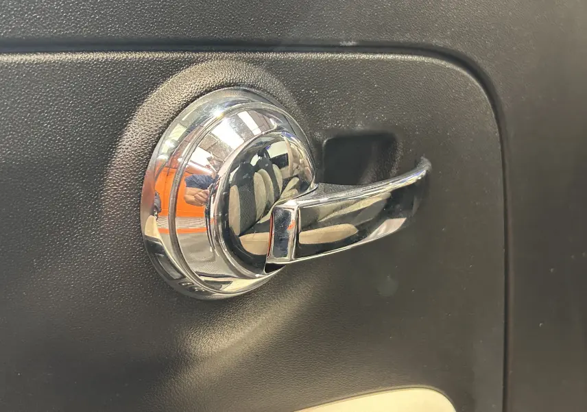 Poignée de porte intérieure chromée sur fond noir, reflet des sièges et de l'habitacle d'une Fiat 500 gris.