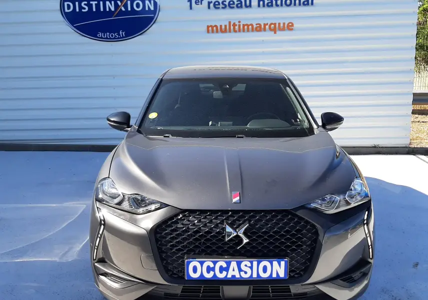 Vue frontale d'un DS3 Crossback gris foncé avec calandre noire et plaque "OCCASION" devant un mur publicitaire.