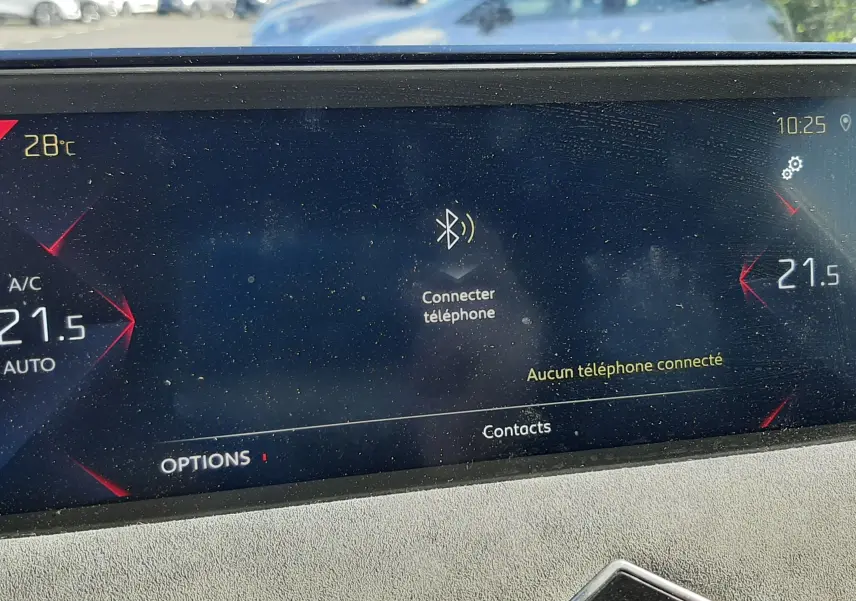 Écran tactile intérieur du DS3 Crossback 2021 affichant la température et la connexion Bluetooth, avec vue floue sur voitures extérieures.
