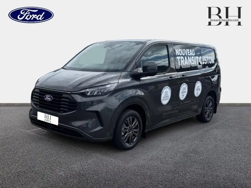 Ford Transit Custom gris métallisé vu en 3/4 avant droit avec marquage publicitaire "Nouveau Transit Custom" sur la porte latérale.