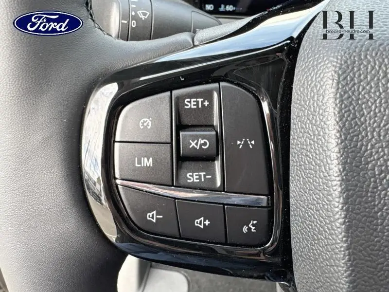 Gros plan sur les commandes au volant noir du Ford Transit Custom Fourgon gris, avec boutons de régulateur et volume.