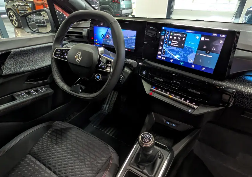 Vue intérieure côté conducteur de la Renault Clio VI TCe 115 Gris Schiste, avec tableau de bord numérique et boîte manuelle.