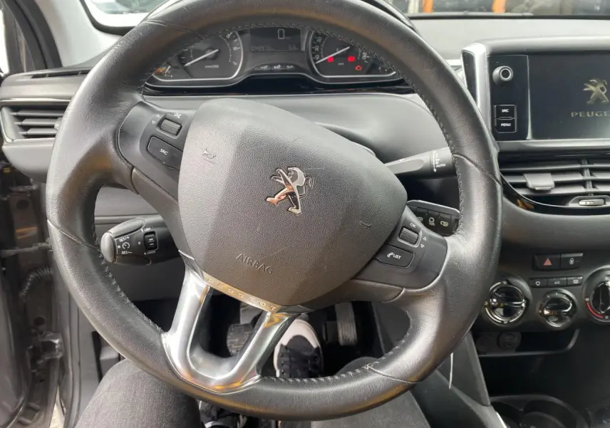 Vue intérieure centrée sur le volant cuir noir du Peugeot 208 gris, avec tableau de bord et écran tactile visibles.