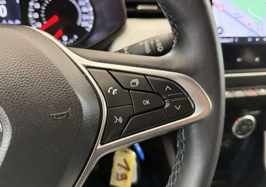 Gros plan sur les commandes au volant cuir noir de la Renault Clio blanche, avec tableau de bord et écran tactile en arrière-plan.