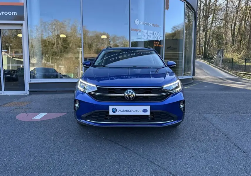 Volkswagen Taigo bleu Recife vue de face, avec calandre noire et phares LED allumés devant showroom.