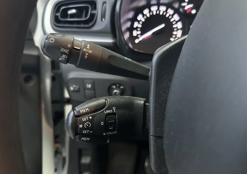 Gros plan sur les commandes au volant côté gauche d'une Citroën C3 blanche, avec tableau de bord flou en arrière-plan.