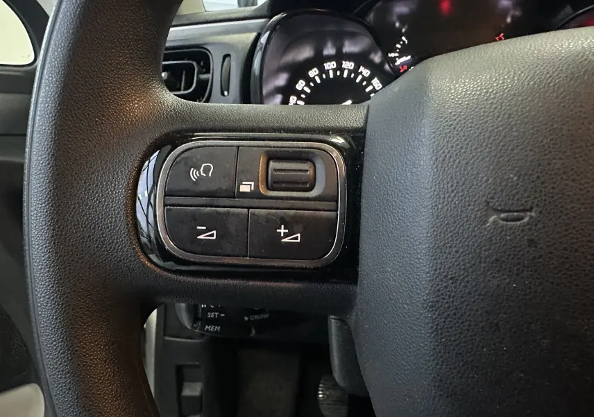 Gros plan sur le volant noir de la Citroën C3 blanc, montrant les commandes audio et le tableau de bord éclairé en arrière-plan.