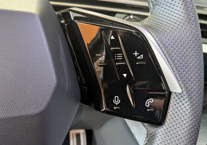 Gros plan sur les commandes noires du volant perforé gris du Peugeot 3008 blanc Okenite, version hybride 2026.