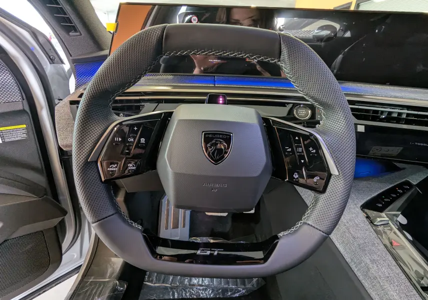 Vue rapprochée du volant GT du Peugeot 3008 blanc Okenite, avec tableau de bord numérique et commandes intégrées.