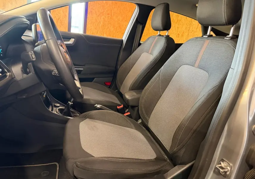 Vue intérieure côté gauche du Ford Puma gris 2021, montrant les sièges avant en tissu noir et gris avec volant et levier de vitesses manuelle.