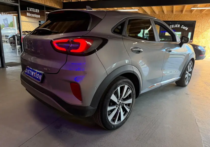 Vue 3/4 arrière droite d'un Ford Puma gris 2021 avec feux arrière allumés dans un garage lumineux.