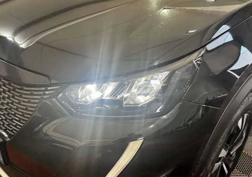 Gros plan sur le phare avant droit allumé d’un Peugeot 2008 noir, mettant en valeur la calandre et la carrosserie brillante.