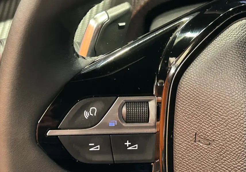 Gros plan sur les commandes vocales et volume au volant noir du Peugeot 2008 II Ph.2 Allure Hybrid 145 bleu Obsession.