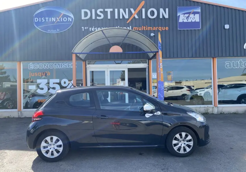 Profil côté gauche d'une Peugeot 208 gris foncé Like 2018 stationnée devant un garage multimarque.