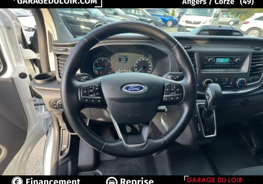 Vue intérieure centrée sur le volant en cuir noir du Ford Transit Custom 2023, avec tableau de bord et levier de vitesses automatique.