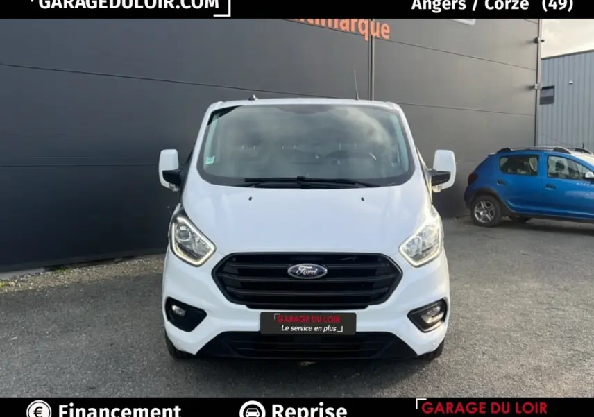 Vue frontale d'un Ford Transit Custom blanc avec calandre noire et phares allumés sur parking extérieur.