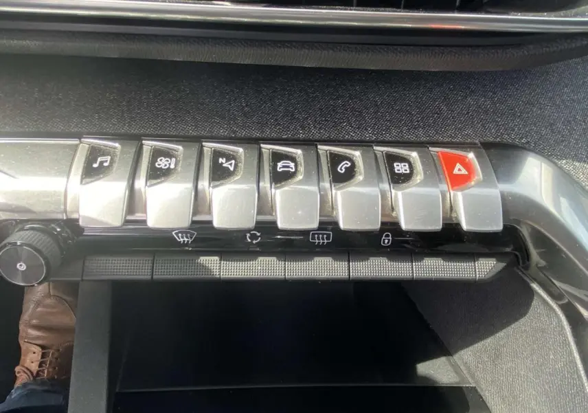 Gros plan sur les commandes en métal brossé du tableau de bord du Peugeot 3008 blanc, avec bouton rouge d'alerte visible.