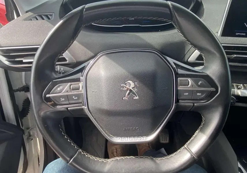 Gros plan sur le volant cuir noir du Peugeot 3008 blanc, mettant en valeur les commandes multifonctions et le logo central.