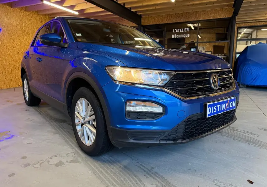 Volkswagen T-Roc bleu en 3/4 avant droit, avec calandre noire et phares allumés en intérieur d'atelier.